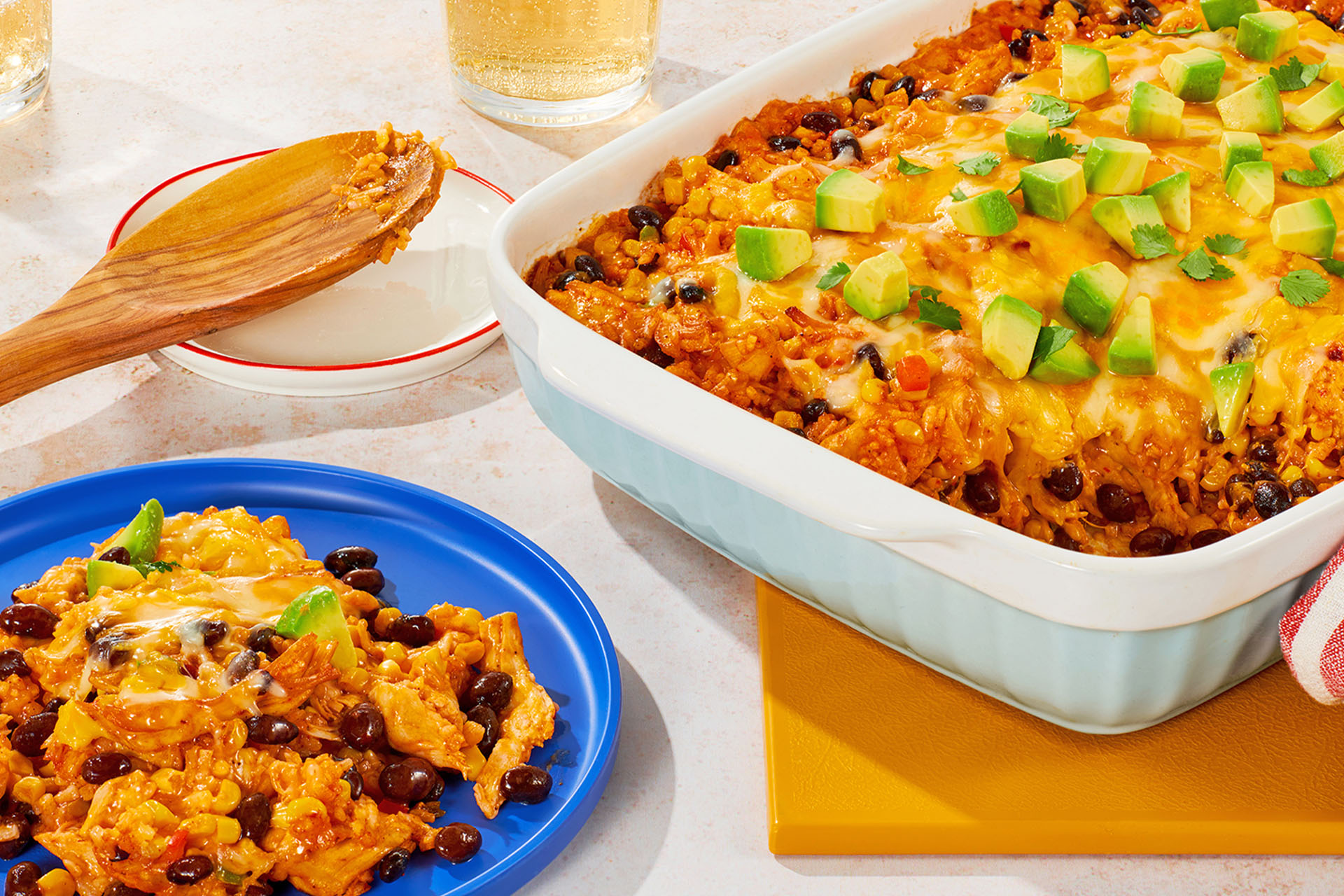 A baked casserole in a white dish topped with avocado and cheese. A serving is dished out on a blue plate.