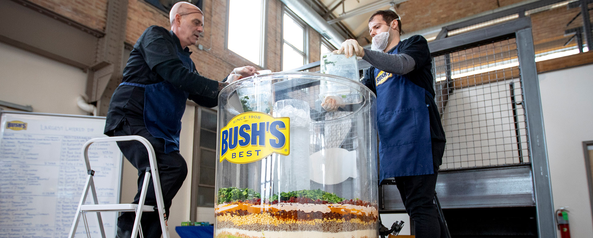 Two men standing on ladders putting ingredients into a large clear cylinder