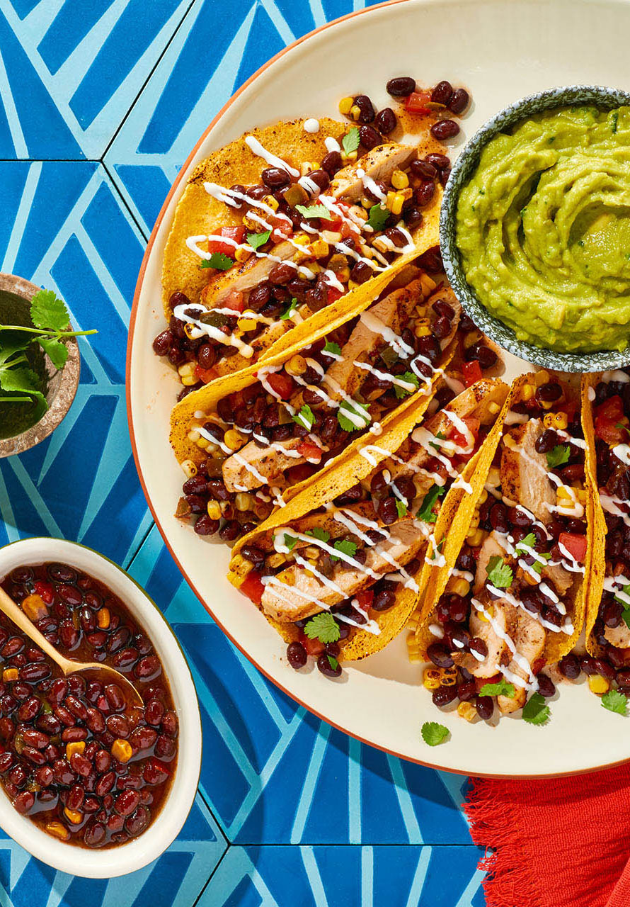 Chicken and black bean tacos with a bowl of guacamole on a blue patterned table.